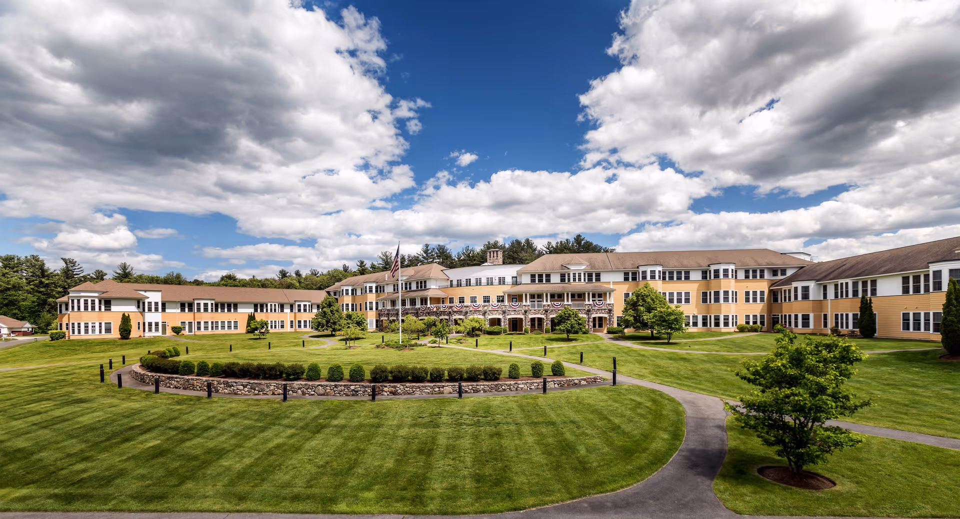 Wide exterior view of Nichols Village senior living facility with a large, well-maintained lawn, a circular garden with shrubs and a flagpole in the center, and a multi-story building with yellow siding and white trim under a partly cloudy blue sky.