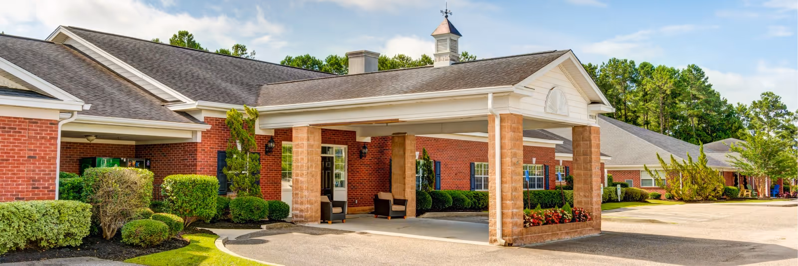 Exterior view of a senior living facility with a covered entrance supported by brick columns, surrounded by well-maintained landscaping including bushes and trees under a partly cloudy sky.