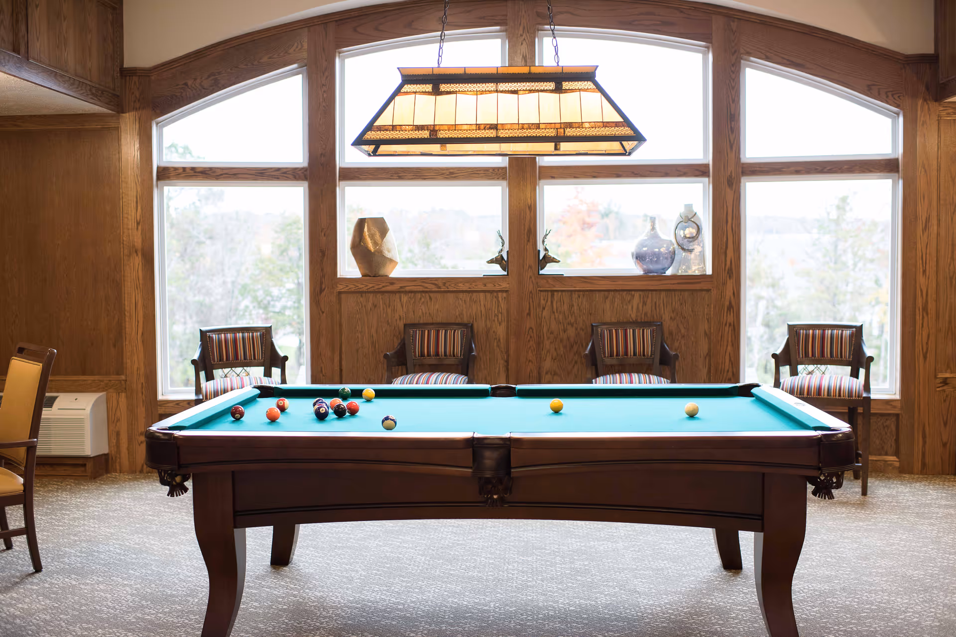A well-lit room with a pool table in the center, several striped cushioned chairs against large windows with wooden frames, and a rectangular hanging light fixture above the pool table.
