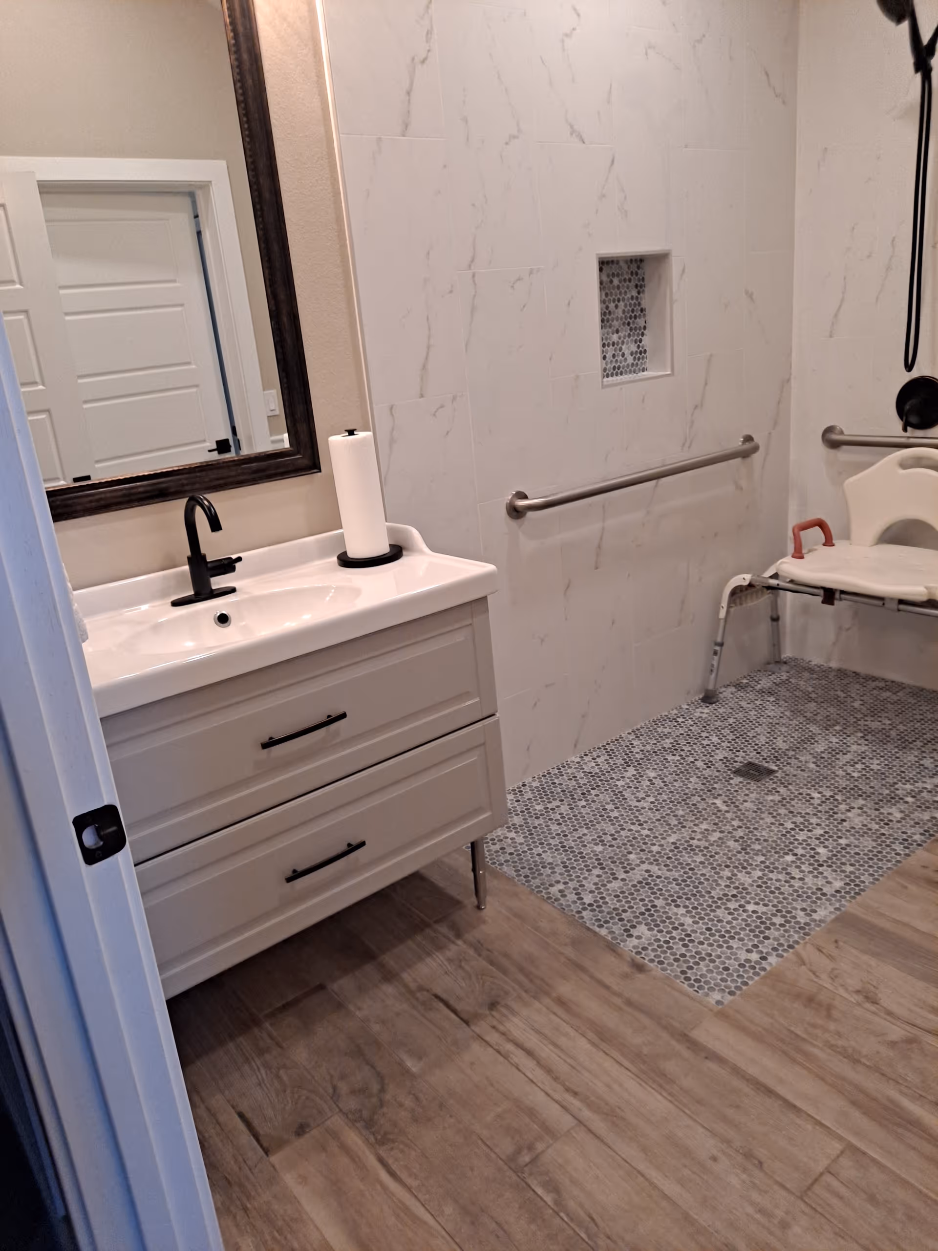 Accessible bathroom with white vanity, mirror, transfer bench, grab bars, and tiled shower.