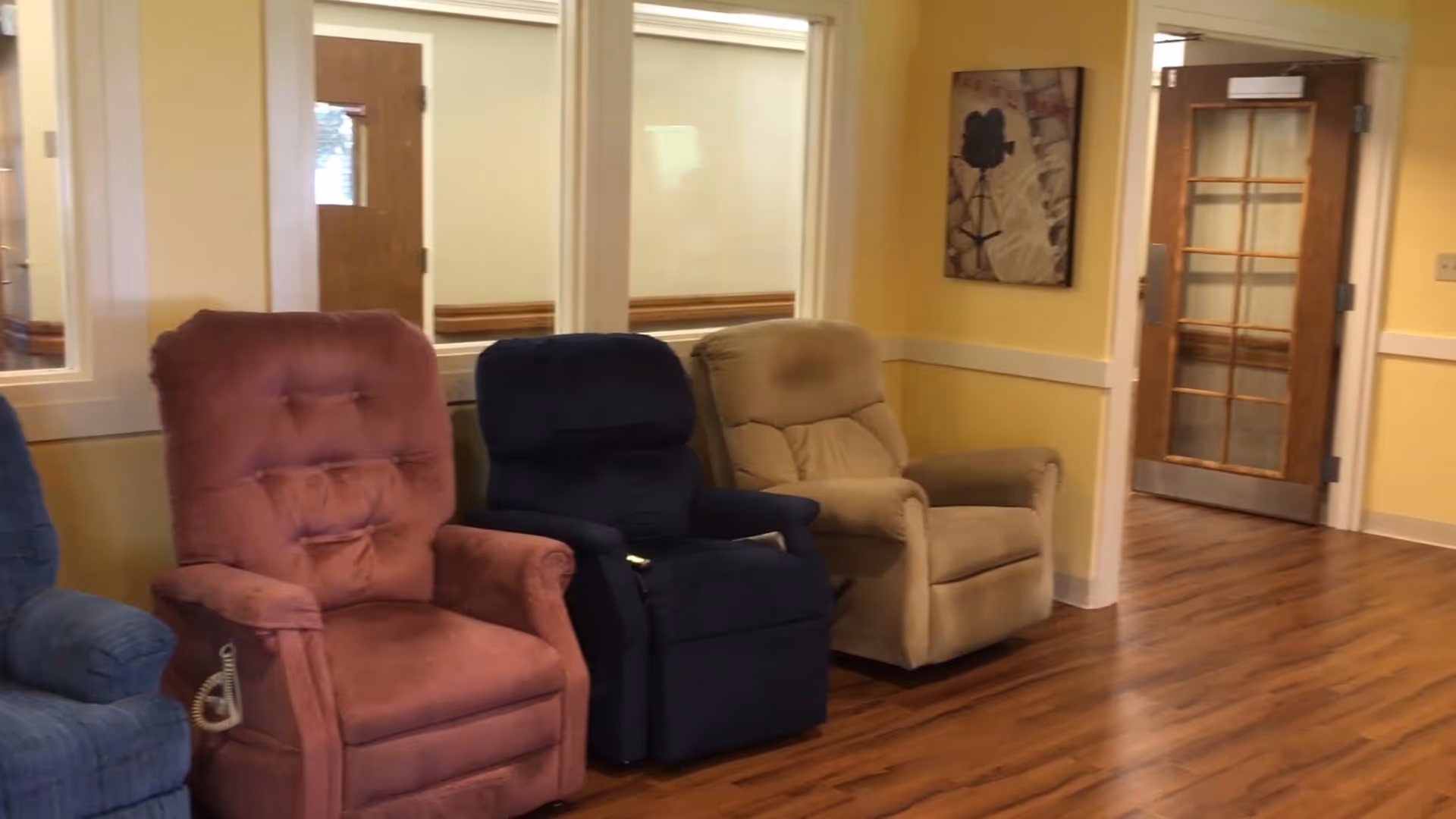 A room with four recliner chairs in different colors (blue, maroon, dark blue, and beige) arranged against a wall with windows. The room has yellow walls, wood flooring, a wooden door with glass panels, and a framed picture on the wall.