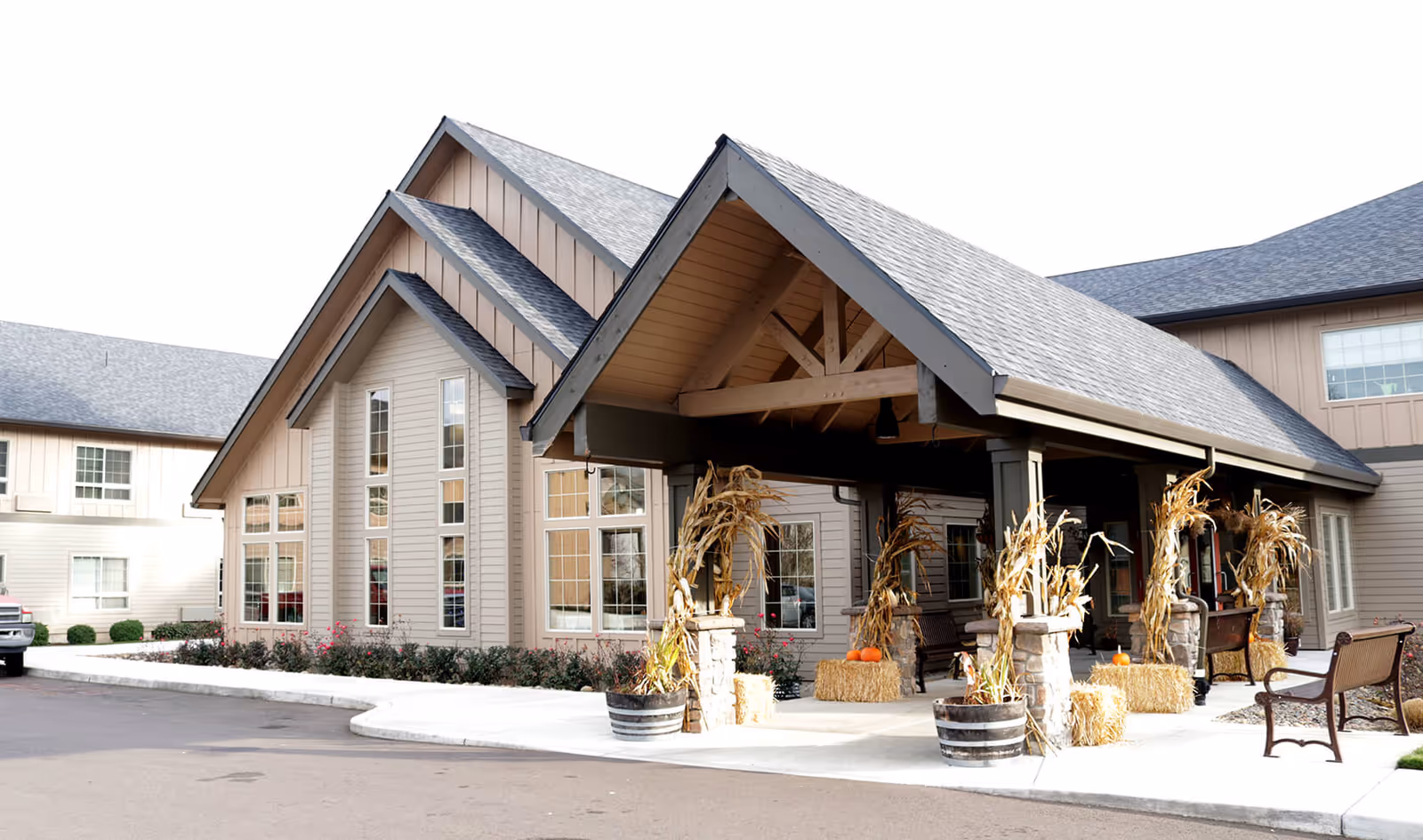 Exterior view of a senior living facility building with a covered entrance decorated with autumn-themed items including hay bales, pumpkins, and dried corn stalks. The building has beige siding, multiple windows, and a gray shingled roof. There is a bench near the entrance and a paved driveway in front.