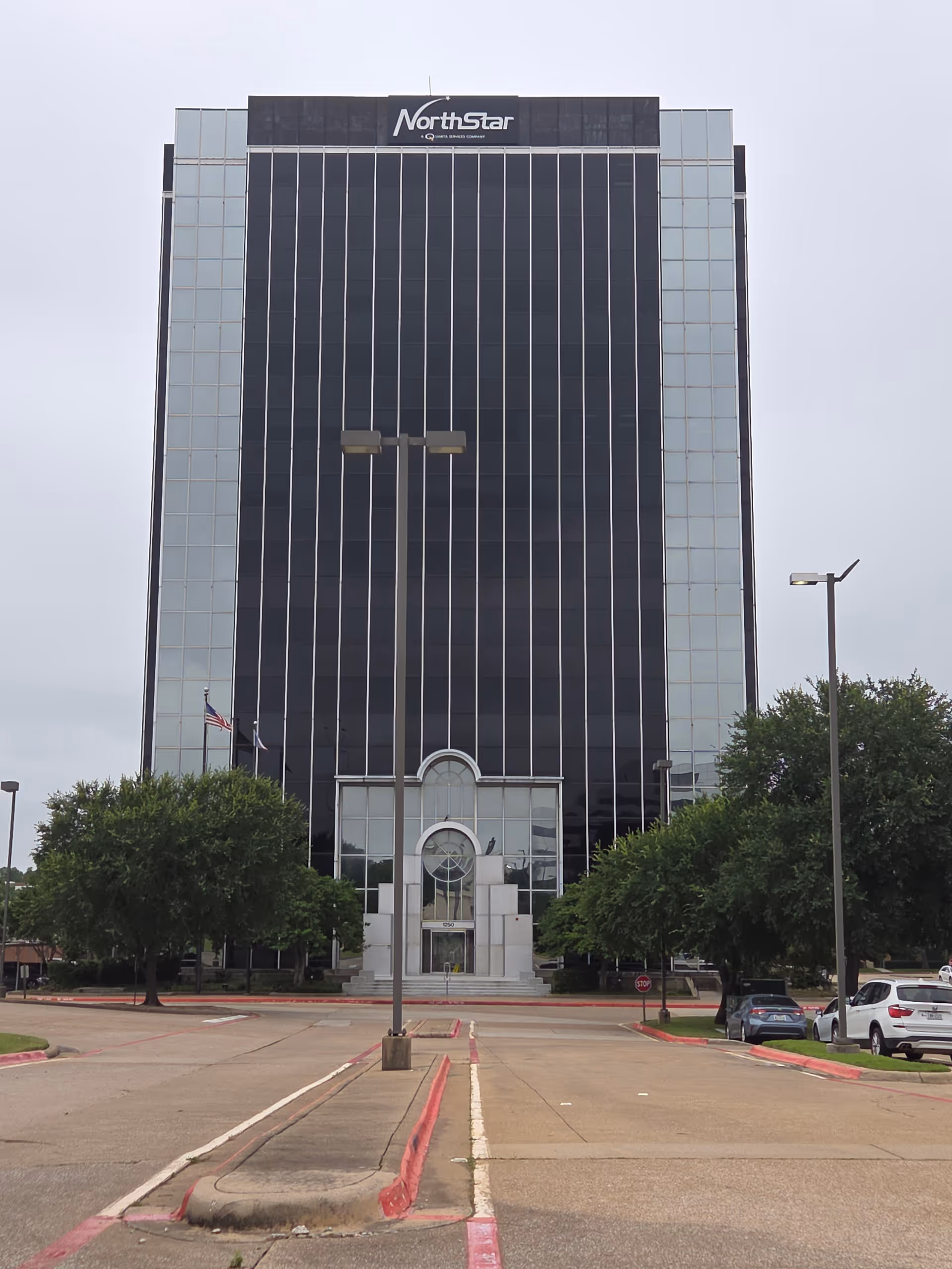 Tall glass-front office building with a central arched entrance and surrounding trees.