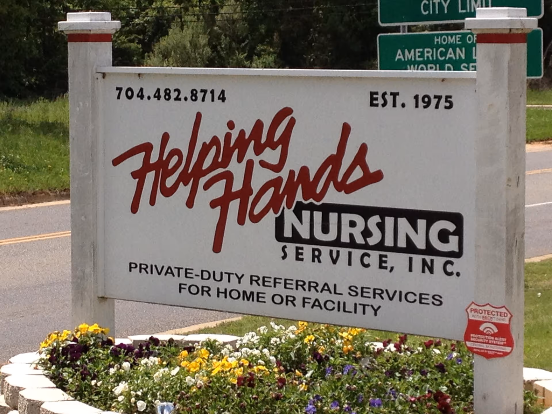 Outdoor sign for Helping Hands Nursing Service with colorful flowers at the base.