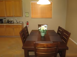 Wood dining table with six chairs and a flower centerpiece in a room with window blinds and cabinets with small appliances.