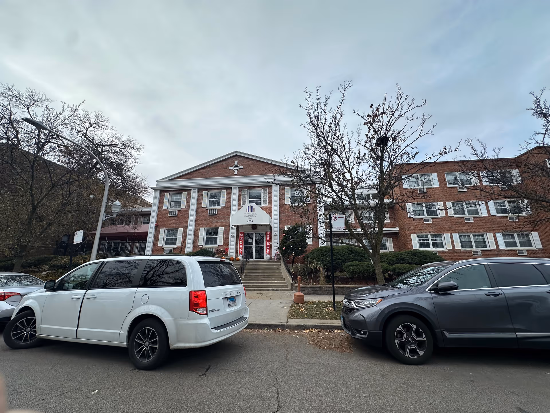 Front view of a three-story brick senior living building with steps leading to the entrance and cars parked along the street.