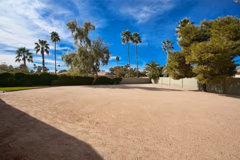 A spacious outdoor area with a sandy ground surface, surrounded by various trees including palm trees and other greenery under a bright blue sky with some clouds.