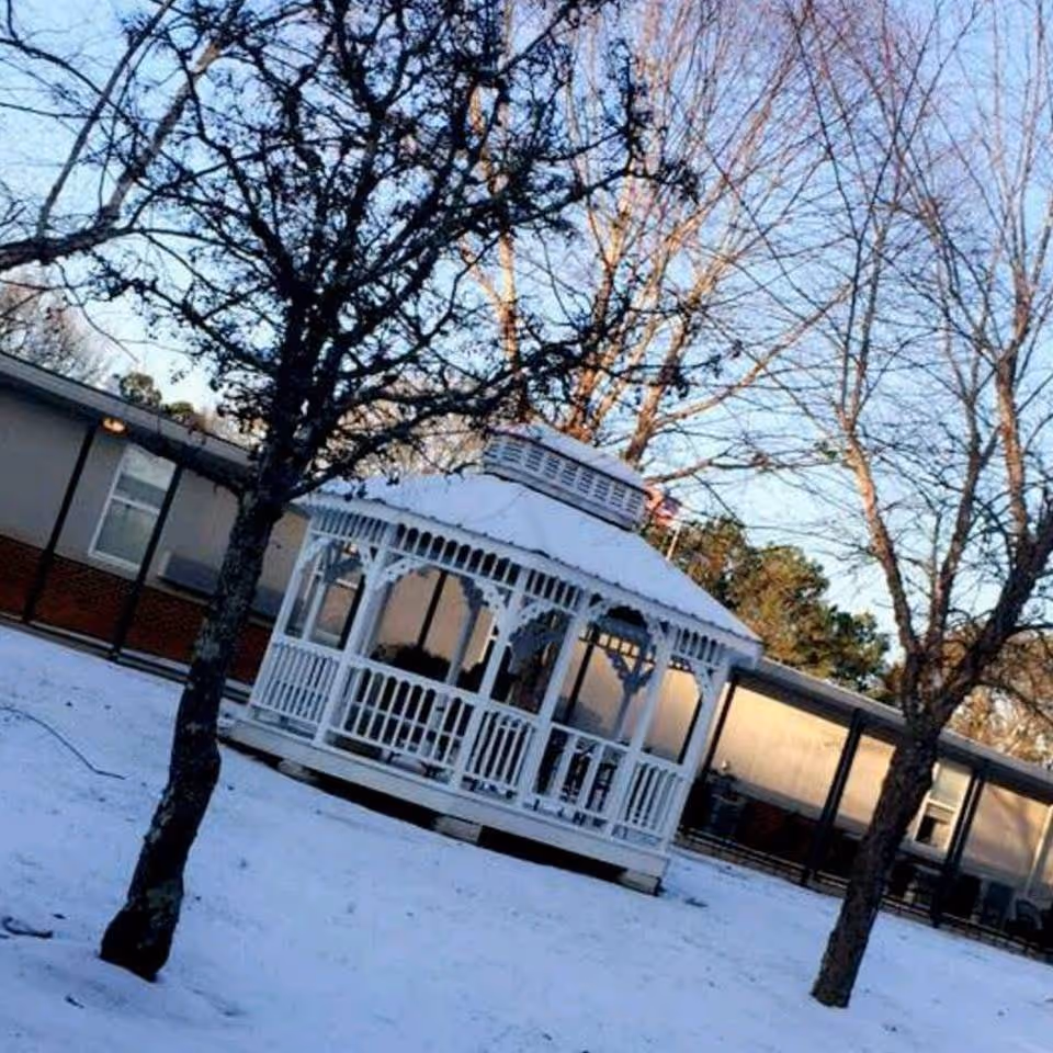 A white wooden gazebo surrounded by snow-covered ground with leafless trees nearby. In the background, there is a single-story building with windows and a covered walkway.