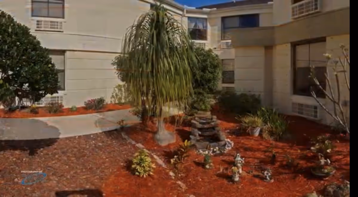 Courtyard garden with a small rock fountain, plants, and a walkway in front of a multi-story senior living building.