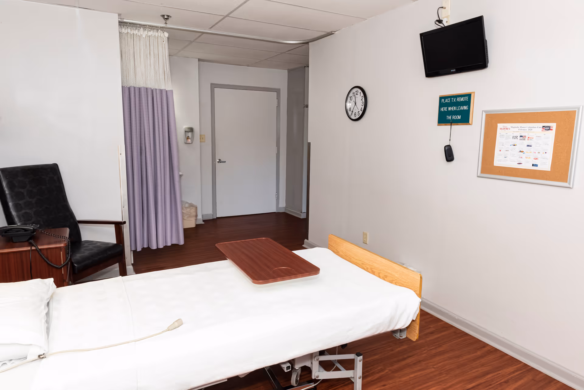 Patient bed with tray, chair, purple curtain, clock, TV, and bulletin board in a room.