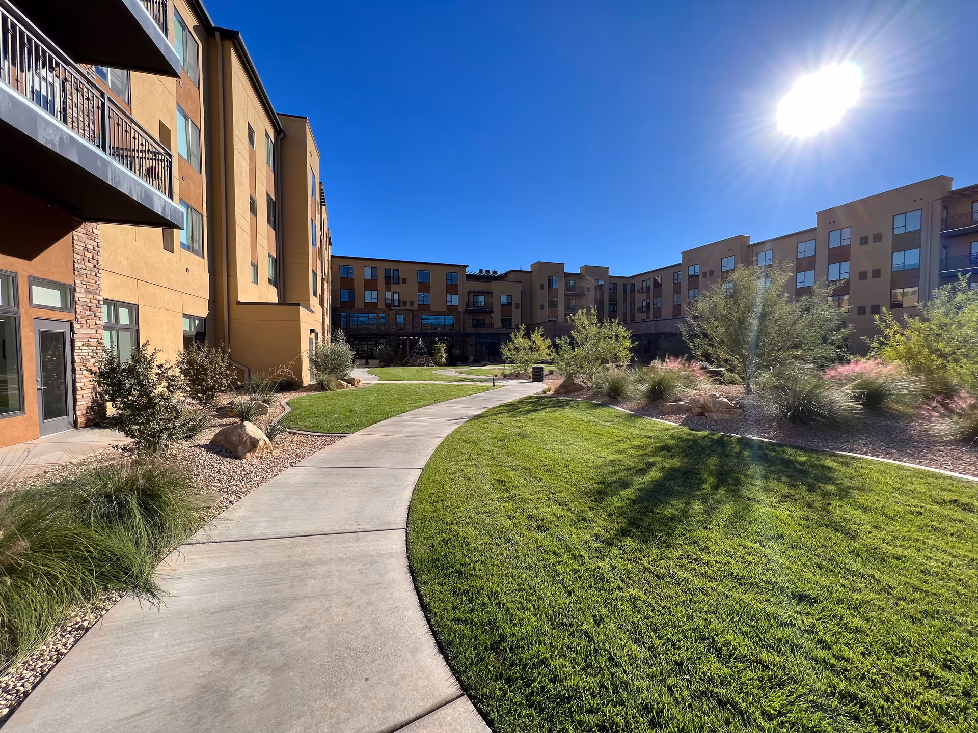 Sunny courtyard with curved walkway, green lawn, and beige apartment buildings surrounding.