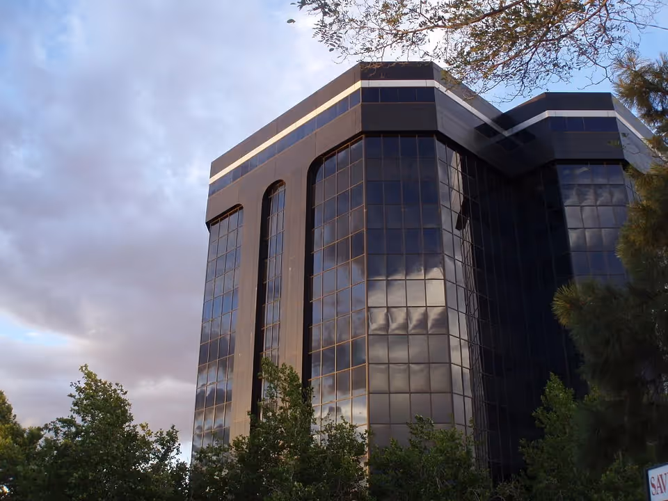Tall glass office building with reflective windows, framed by trees and cloudy sky.