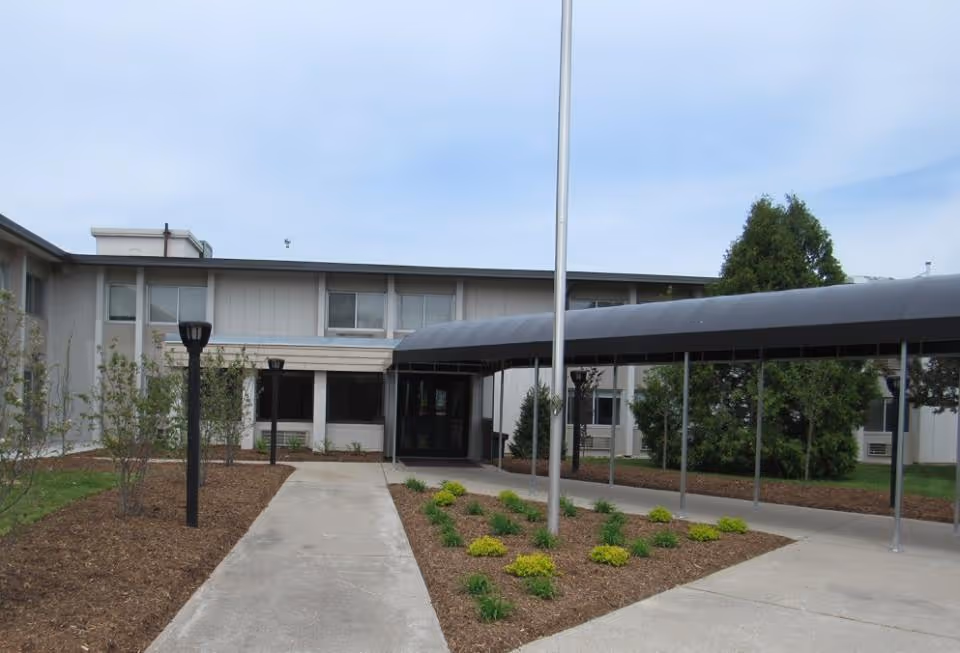 Exterior view of Dimondale Nursing & Rehab Center showing a two-story building with a covered walkway leading to the entrance, surrounded by landscaped areas with small bushes and trees under a partly cloudy sky.
