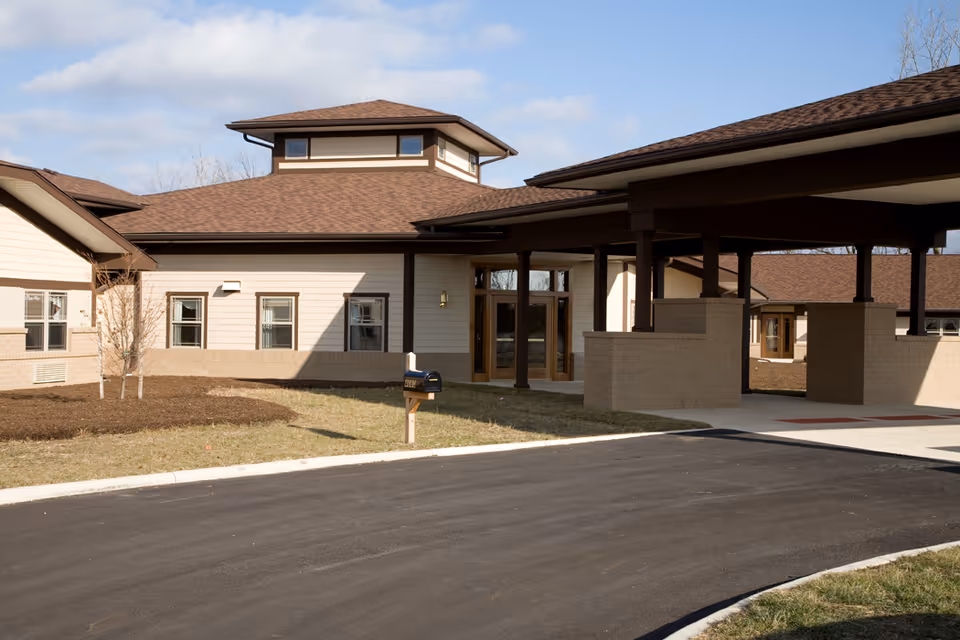 Exterior view of a single-story senior living facility building with beige siding and brown roof shingles. The building features multiple windows, a covered entrance supported by columns, and a paved driveway leading up to the entrance. The sky is partly cloudy.