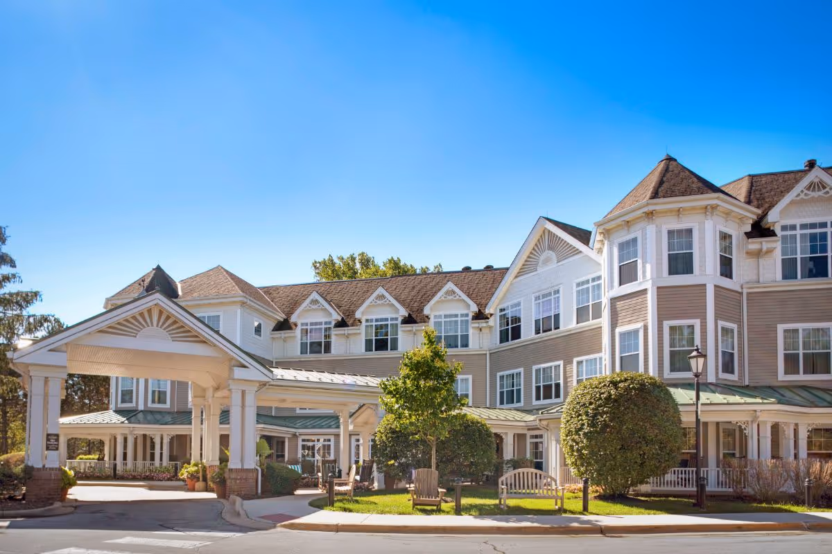 Front exterior of a multi-story senior living building with a covered entrance, manicured shrubs, and benches under a clear blue sky.