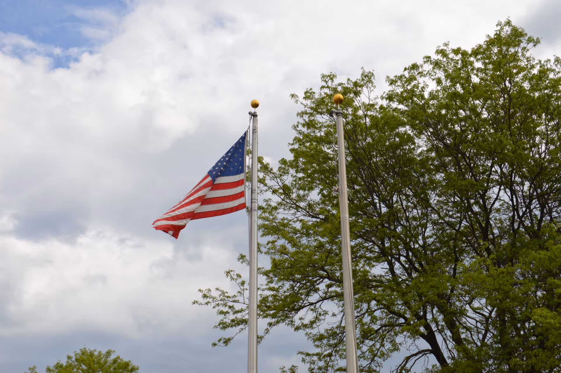 An American flag flying on a flagpole beside another pole with trees and a cloudy sky in the background.