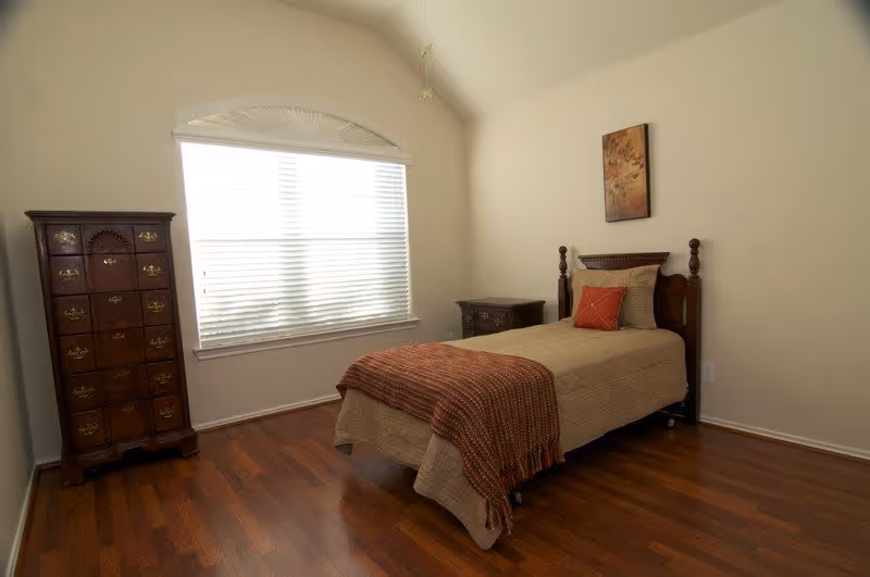 A simple bedroom with a single bed covered in beige bedding and a rust-colored throw blanket and pillow. The room has wooden flooring, a tall wooden dresser on the left, a small wooden nightstand on the right, and a large window with white blinds letting in natural light. A framed floral artwork hangs above the bed on the wall.