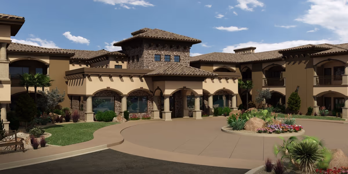 Exterior view of The Retreat at Sunriver senior living facility showing a large, multi-story building with a tiled roof, stone and stucco facade, arched windows and doorways, surrounded by landscaped greenery and a circular driveway with a flower bed in the center under a partly cloudy blue sky.