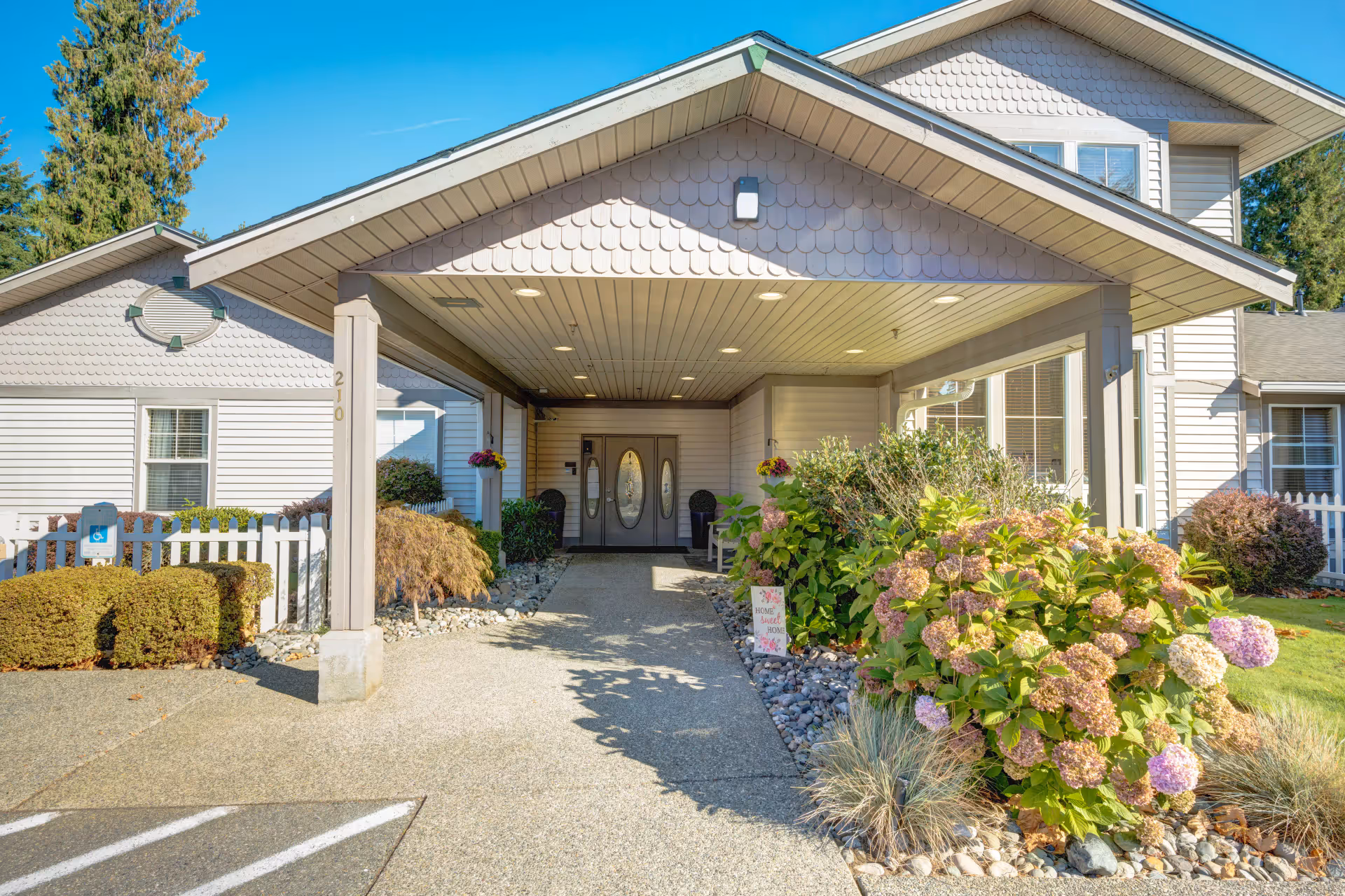 Covered entrance with double glass doors, a paved walkway and flowering shrubs at the front of a residential care building.