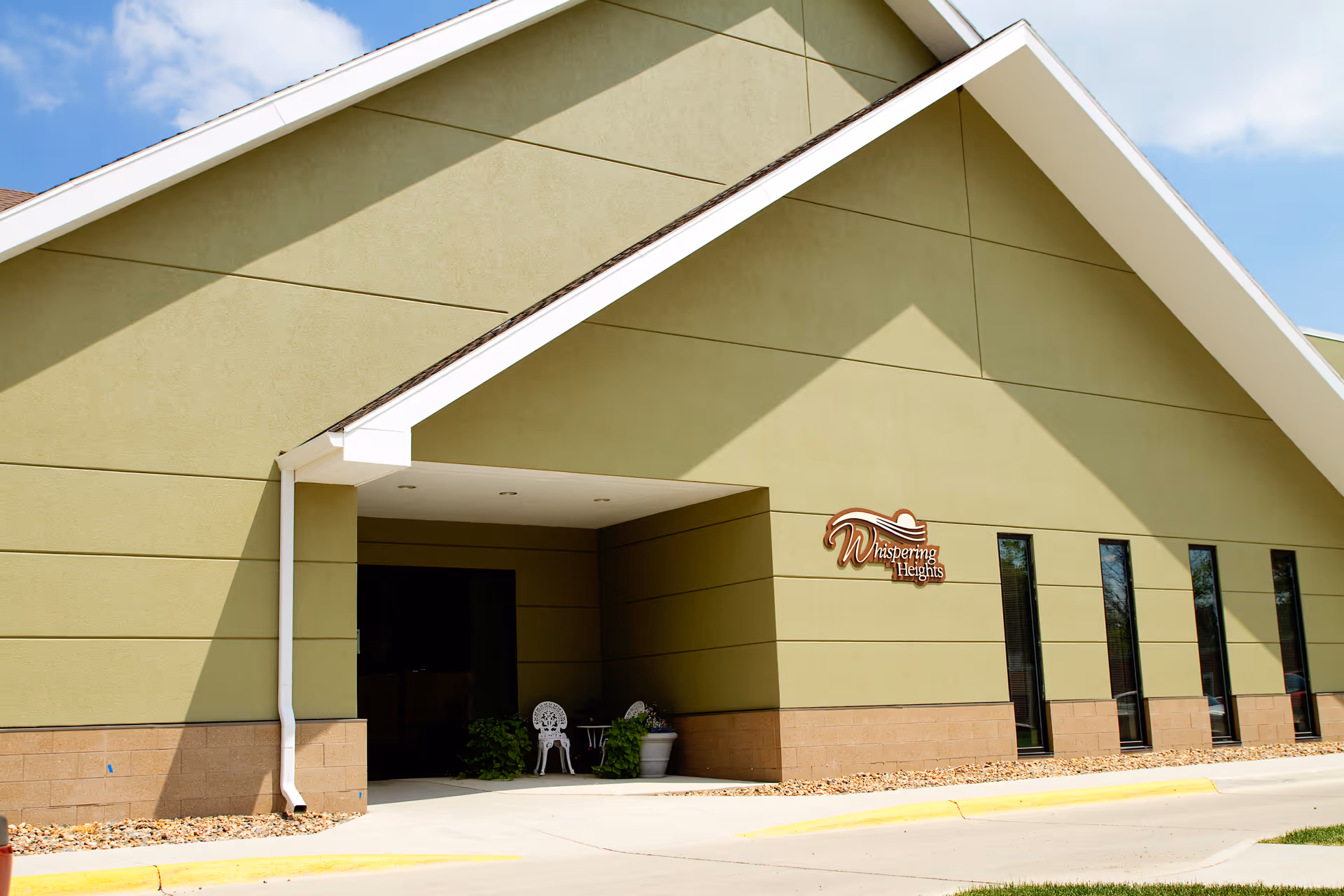 Exterior view of a building with a green and beige facade under a blue sky. The building has a covered entrance with two white chairs and a small table, along with some potted plants. A sign on the wall reads 'Whispering Heights'.