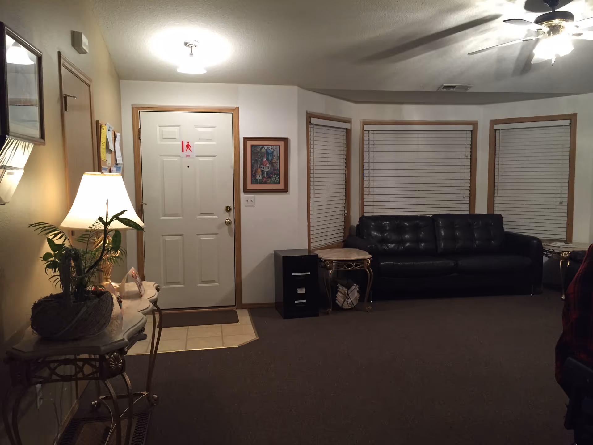 Living room interior of a senior care home with a black leather couch, lamp, and blinds.