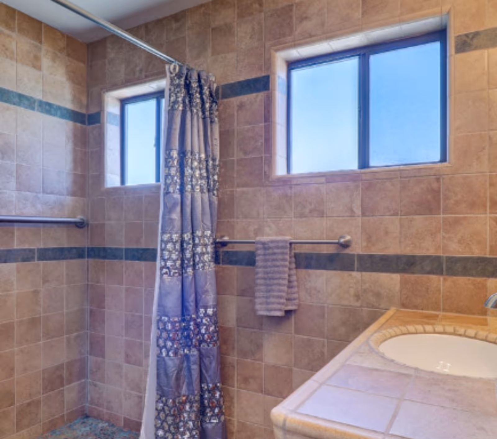 Tile bathroom with shower curtain, towel rack, two windows, and sink.