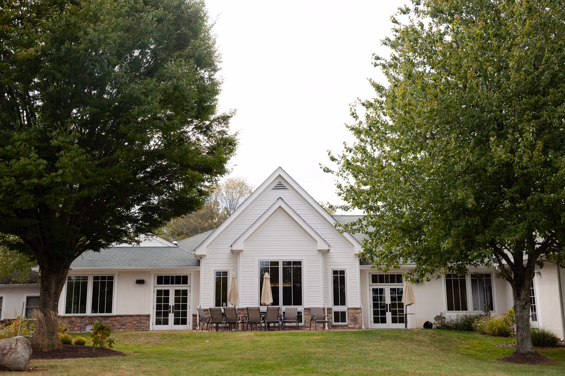 Front exterior of a white single-story building with a patio area, tables and umbrellas, flanked by large trees and a grassy lawn.