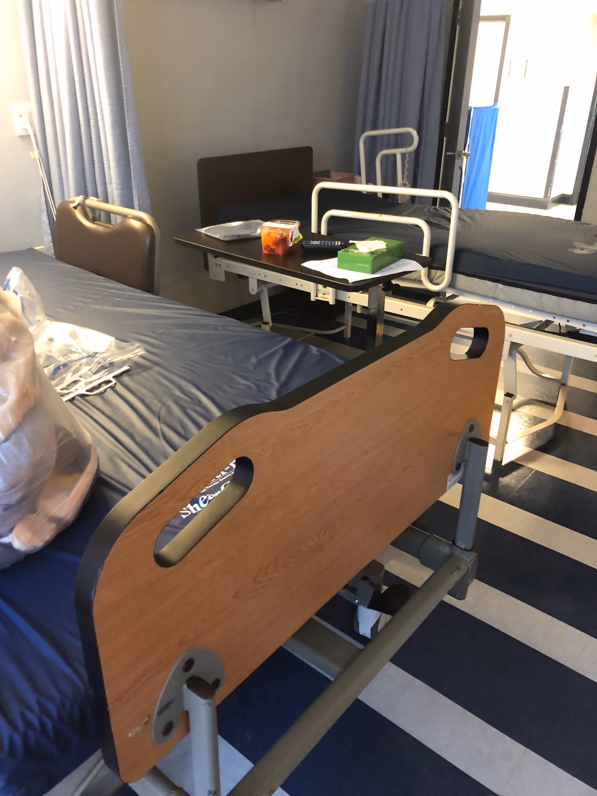 Interior hospital-like patient room with adjustable beds, blue curtains, and a bedside table.