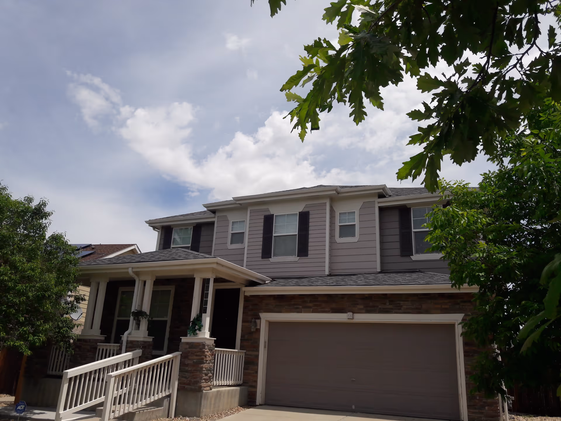 Exterior view of a two-story residential building with a garage, a front porch with a ramp, and surrounded by trees under a partly cloudy sky.
