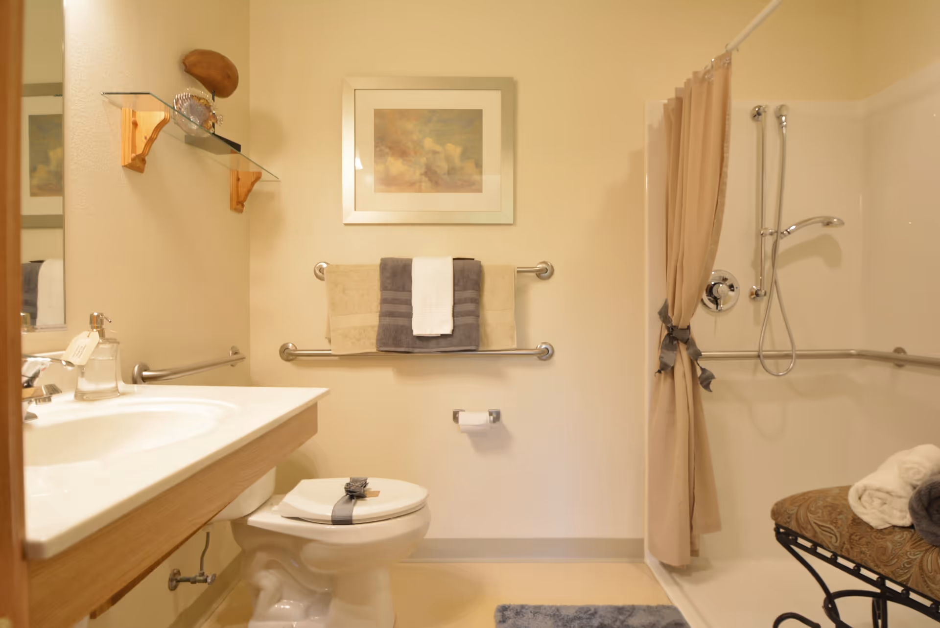A clean and well-lit bathroom featuring a white toilet with a decorative ribbon, a sink with a soap dispenser, a towel rack with beige, gray, and white towels, a shower with a beige curtain tied back, and a cushioned bench with rolled towels. A framed picture hangs above the towel rack and a glass shelf with decorative items is mounted on the wall.