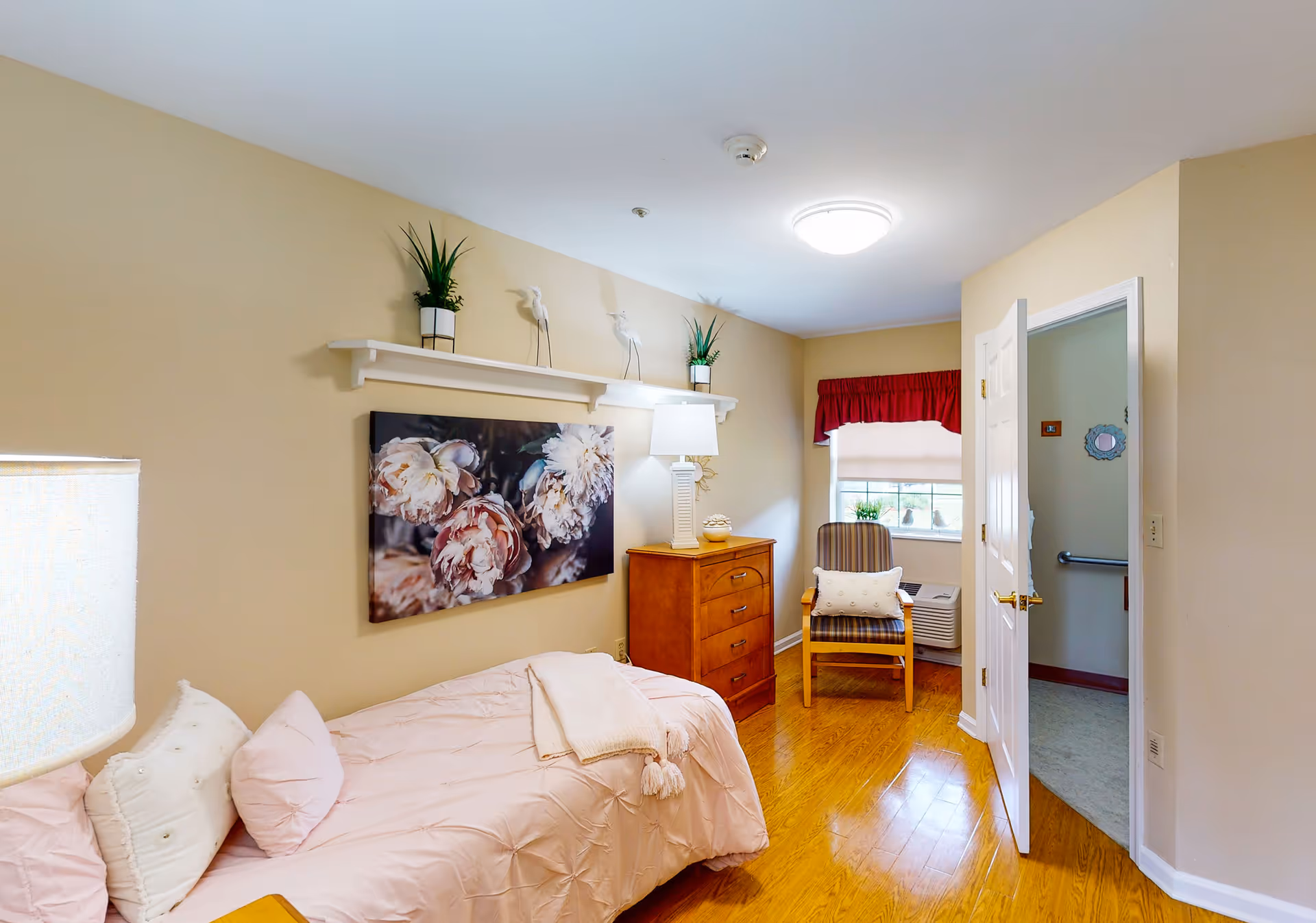 Bedroom in a senior living facility with pink bedding, dresser, chair, and window.