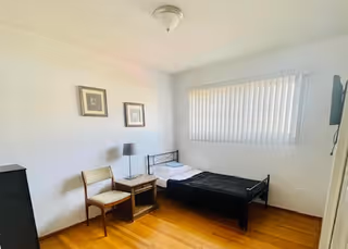 Small bedroom with a single bed, chair, nightstand, lamp, and window blinds.
