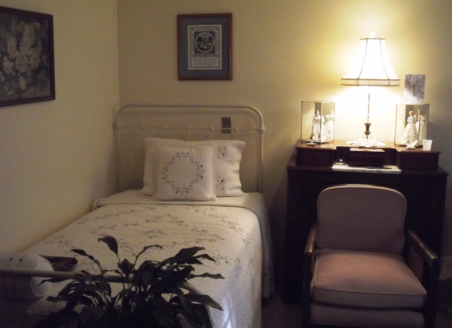 A cozy bedroom with a single bed covered in a white quilt and matching pillows. Next to the bed is a wooden desk with a lit table lamp and decorative figurines in glass cases. A cushioned armchair is placed in front of the desk. The walls have framed artwork, and a potted plant is visible in the foreground.