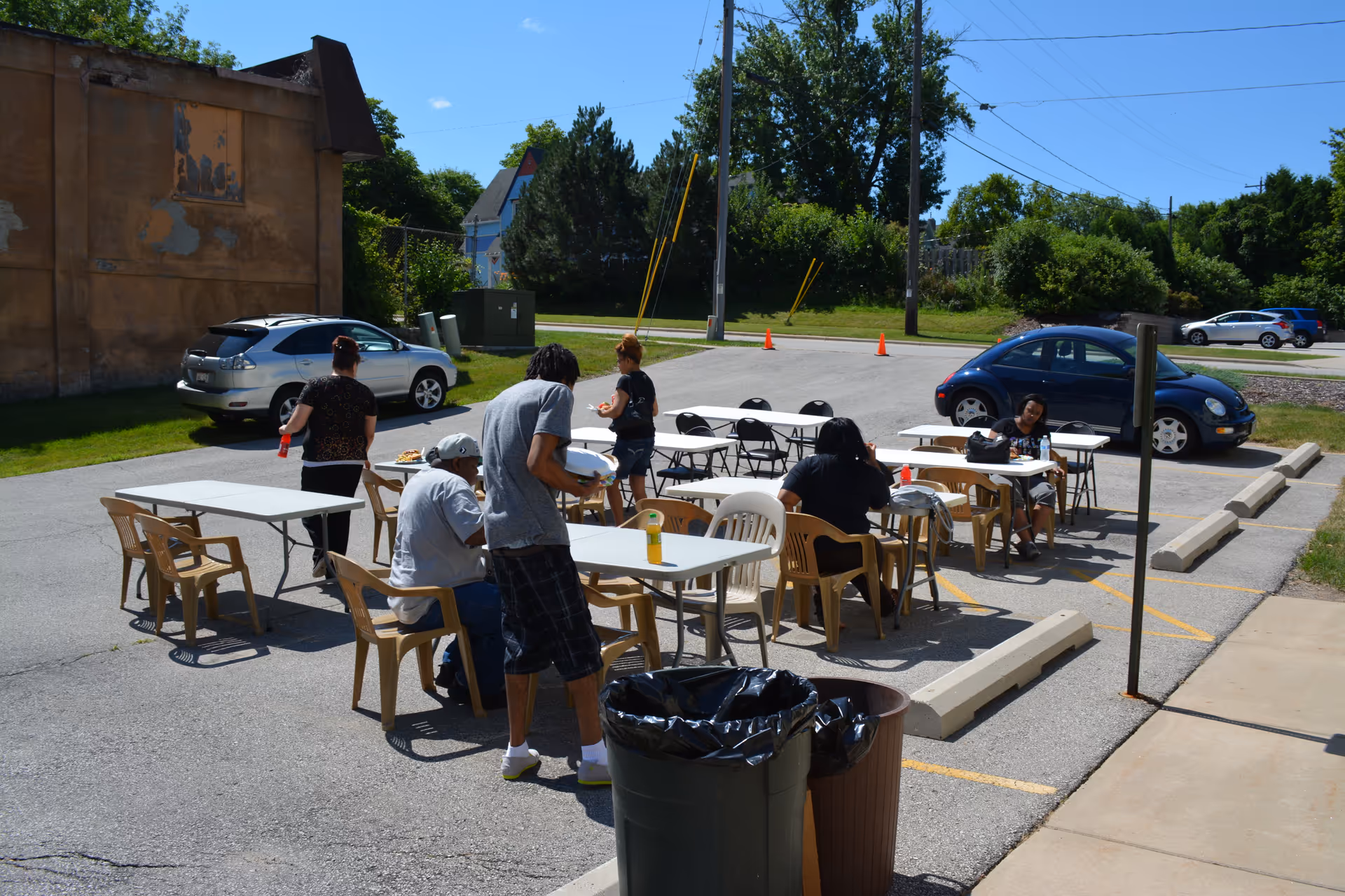 Outdoor parking lot with picnic tables, plastic chairs, and people gathered.