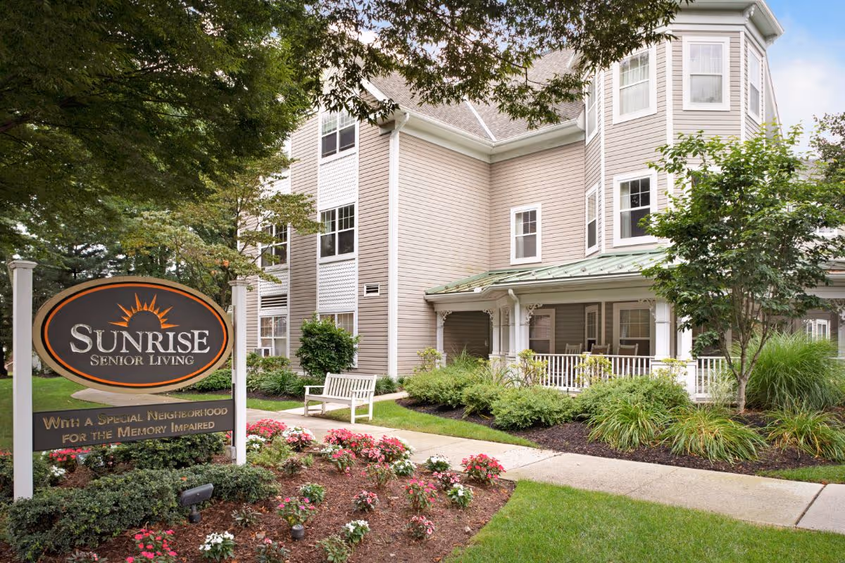 Front exterior of the Sunrise Senior Living building with a large sign, porch, bench, and landscaped flowerbeds.