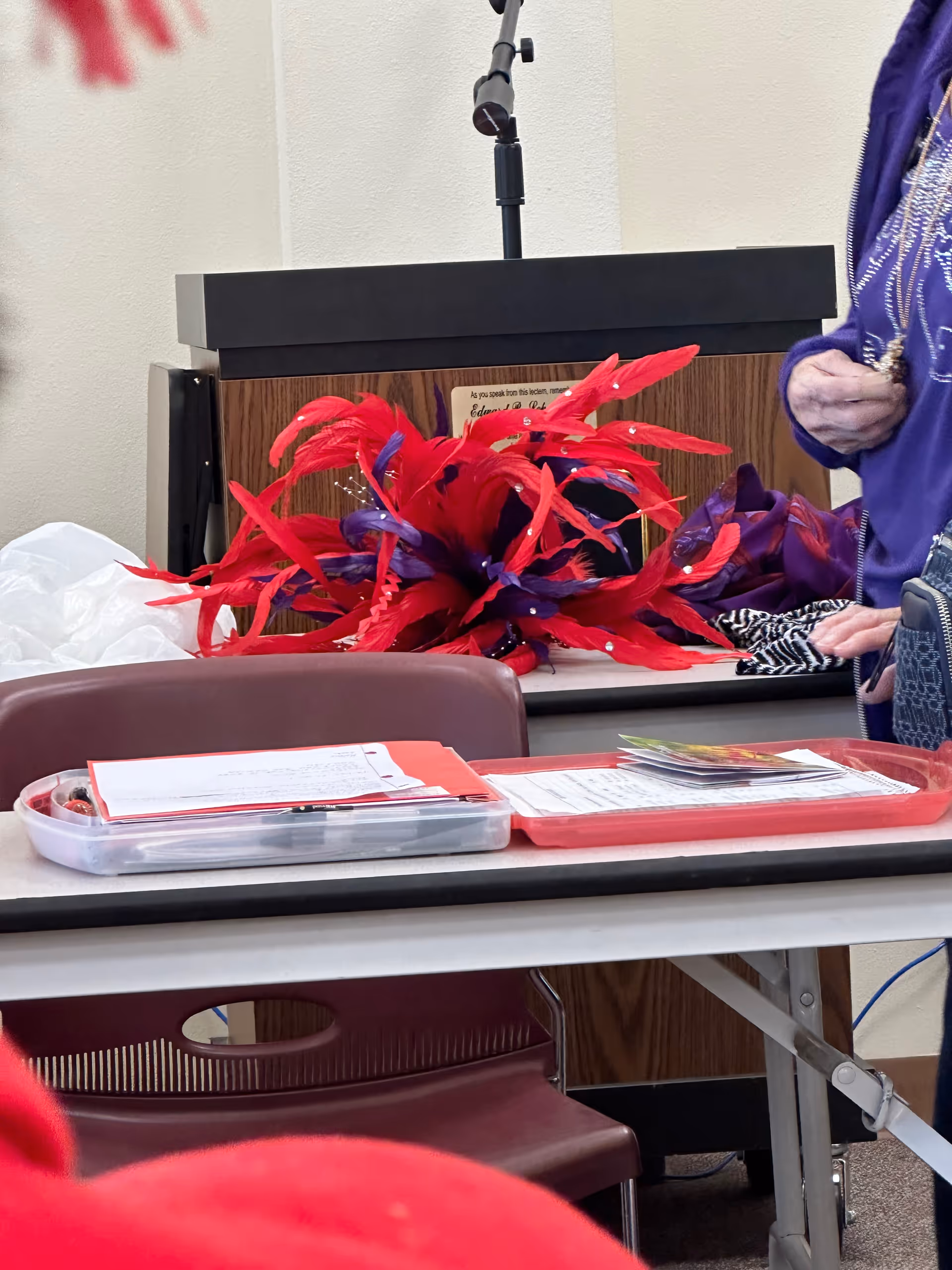 Interior meeting space with a wooden podium, microphone, and red-purple feather centerpiece.