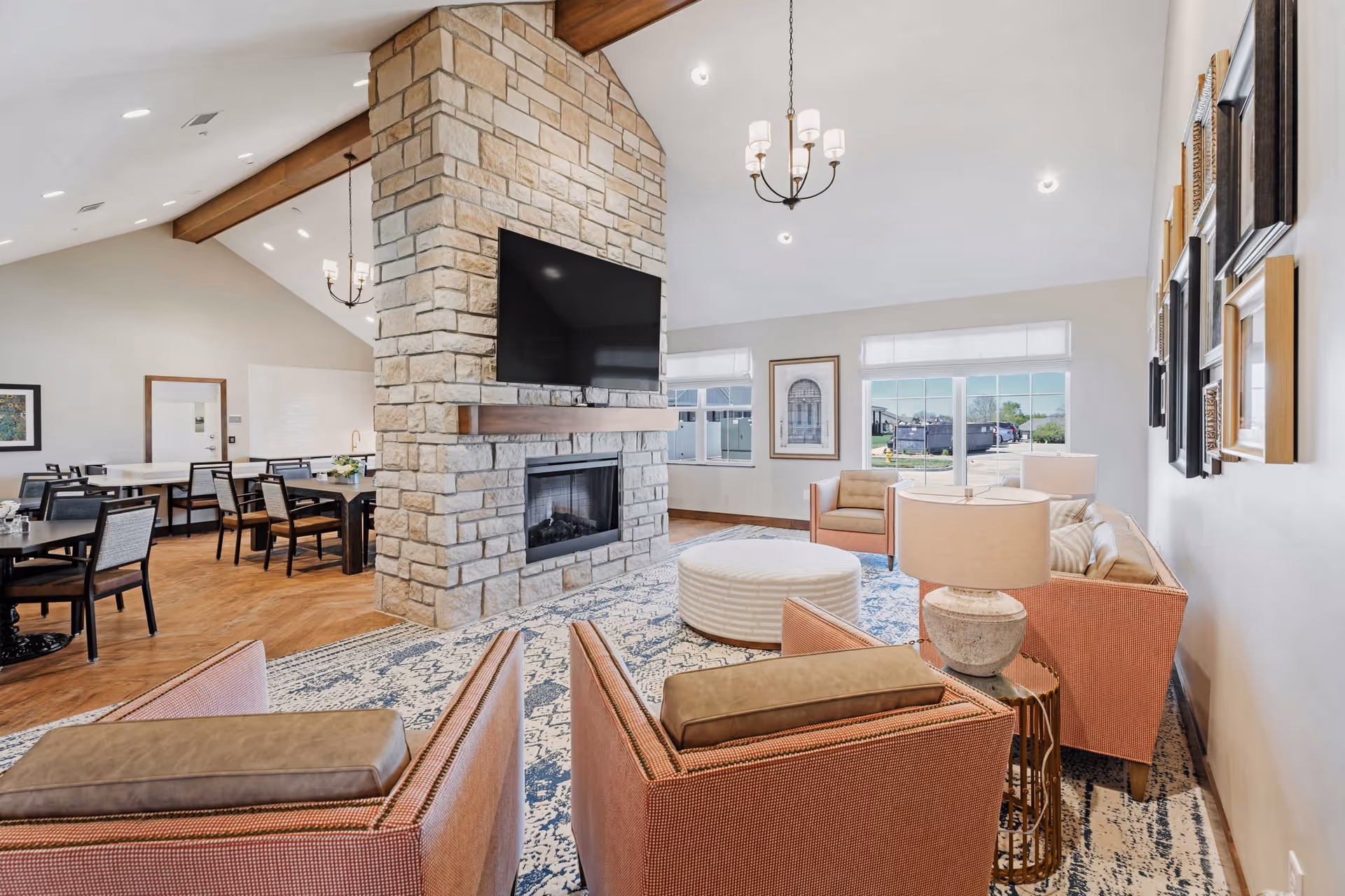 A bright and spacious senior living common area featuring a stone fireplace with a mounted flat-screen TV. The room has comfortable seating including armchairs and a sofa arranged around a round ottoman on a patterned rug. In the background, there are dining tables and chairs, large windows letting in natural light, and framed artwork on the walls.