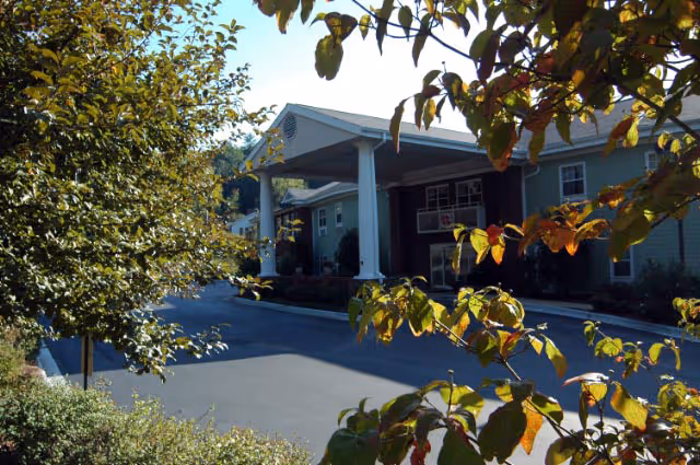 Entrance of a two-story building with white columns and a driveway framed by trees and foliage.