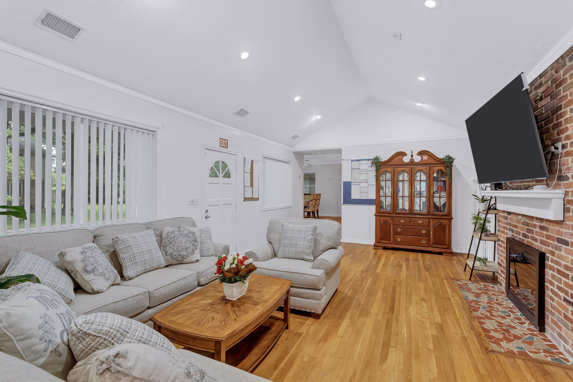 Spacious living room with beige sectional sofa, wooden furniture, brick fireplace, TV, and hardwood floors.