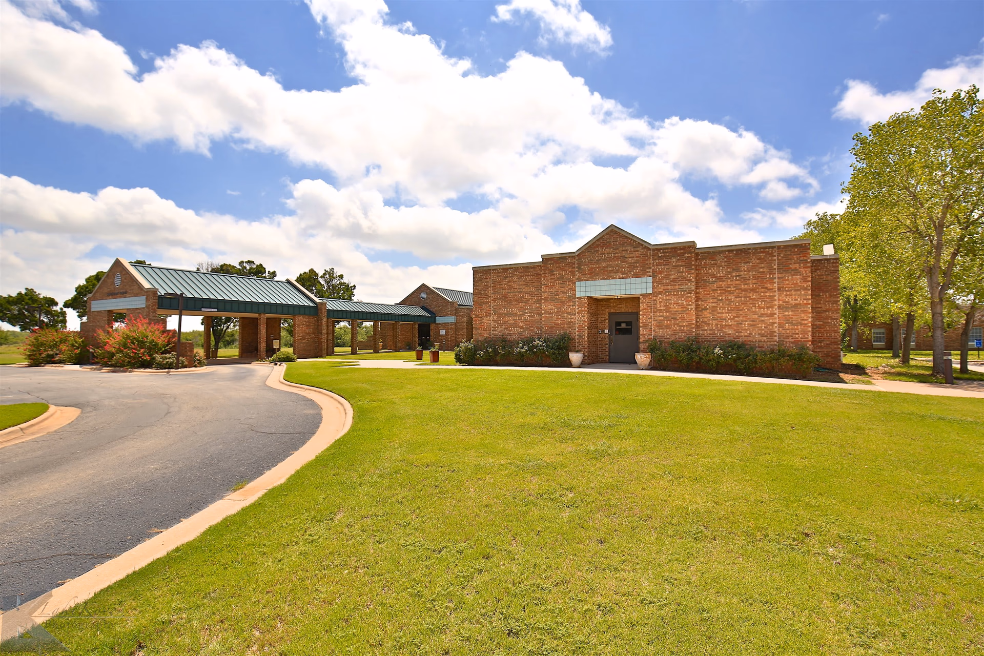 Exterior view of Mesa Springs Healthcare Center showing a brick building with a covered entrance and a driveway surrounded by green grass and trees under a partly cloudy blue sky.