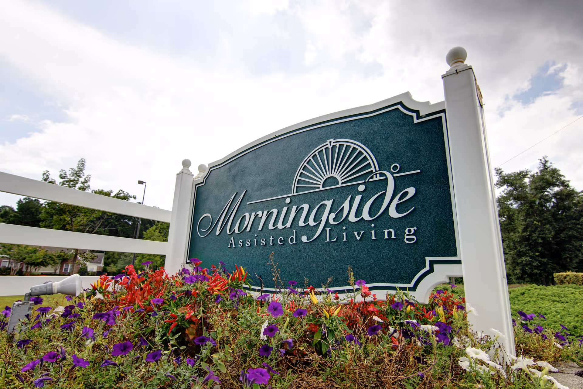A large green and white sign for Morningside Assisted Living surrounded by colorful flowers and greenery, with a white fence and trees in the background under a partly cloudy sky.