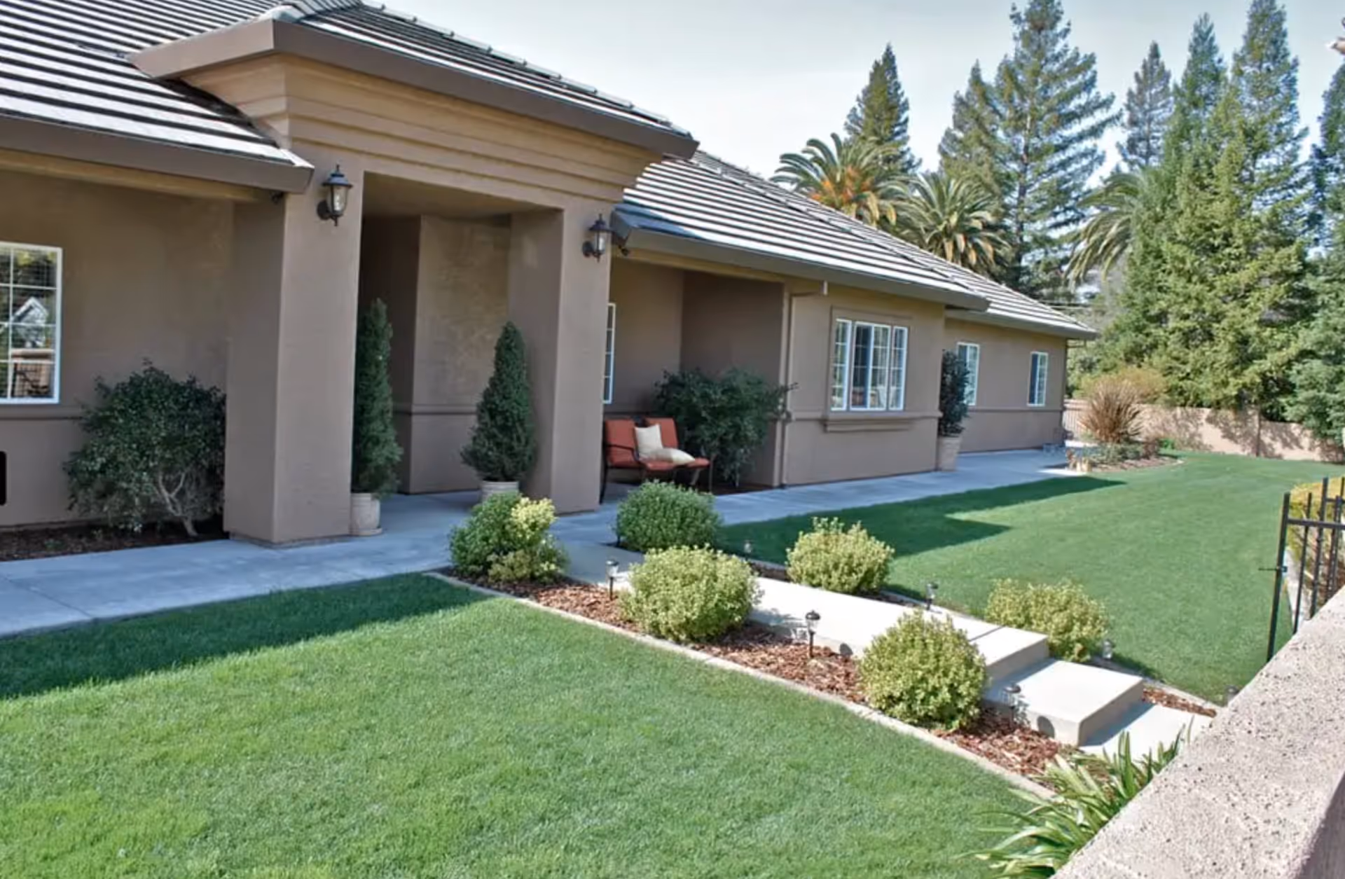 Exterior view of a single-story building with a covered entrance, beige walls, and a tiled roof. The building is surrounded by a well-maintained lawn, small shrubs, and trees, with a concrete pathway leading to the entrance. There is a red cushioned bench with pillows near the entrance.