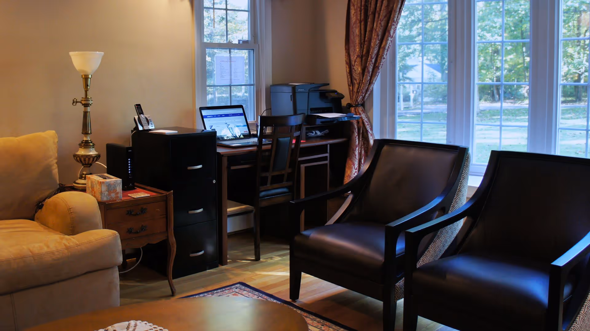 A cozy interior room with a beige armchair, a wooden side table with a lamp and tissue box, a black filing cabinet, a wooden desk with a laptop and printer, two black leather armchairs, and large windows with patterned curtains letting in natural light.