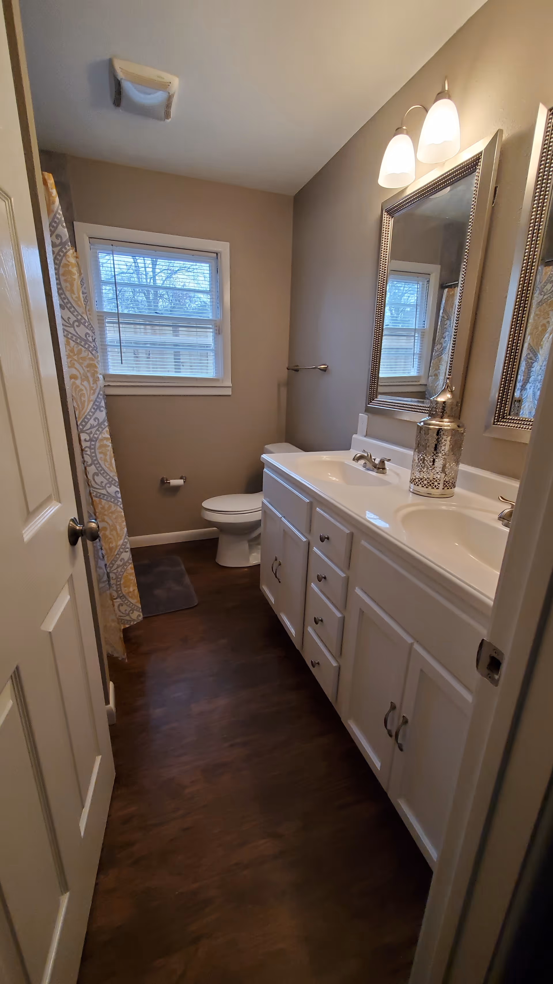 Narrow bathroom with double vanity, toilet, window, shower curtain, and modern lighting.