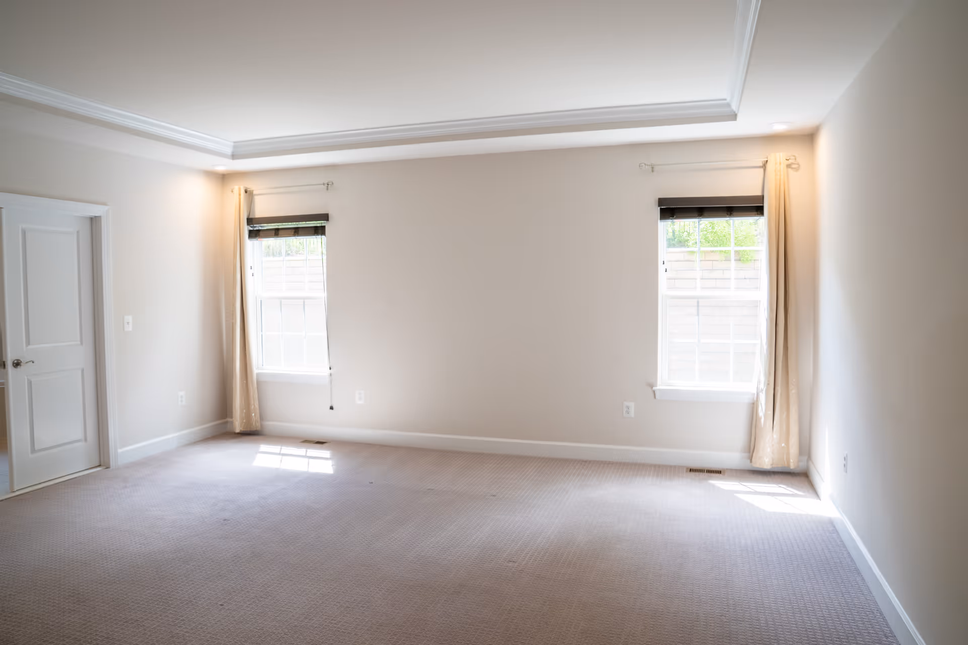 Bright empty living room with two windows, beige walls, curtains, and carpet.