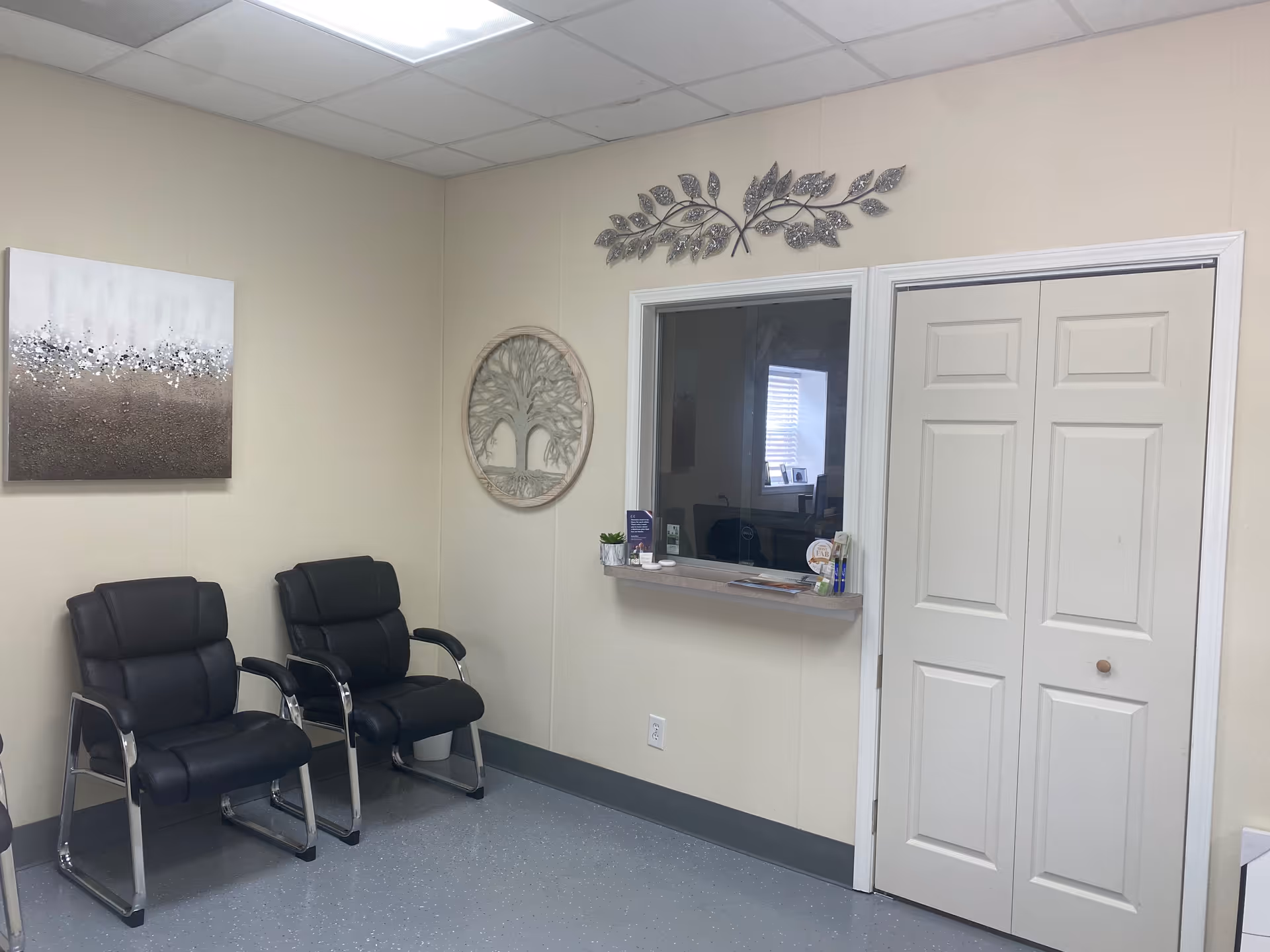 A small beige waiting area with black chairs, wall art, and a windowed reception shelf.