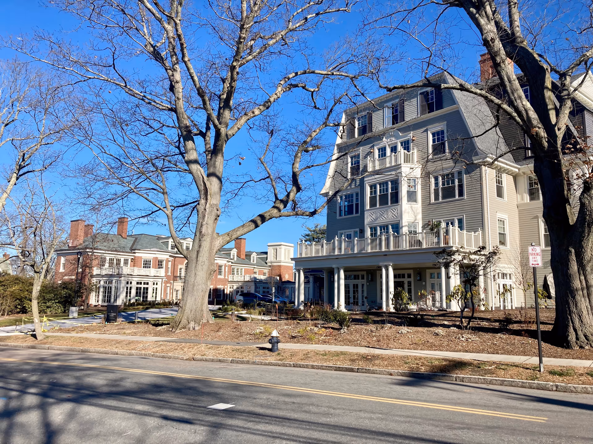 Exterior view of The Newbury Of Brookline senior living facility showing a large multi-story building with numerous windows and balconies, surrounded by leafless trees and a clear blue sky.