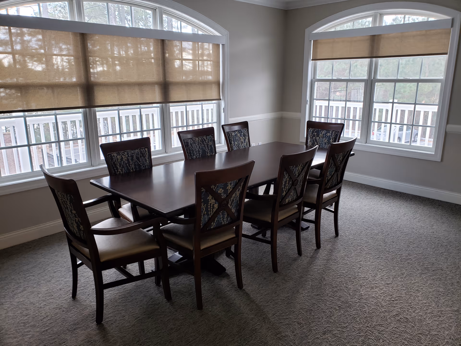 A long dining table with eight chairs in a bright, window-lit dining room.