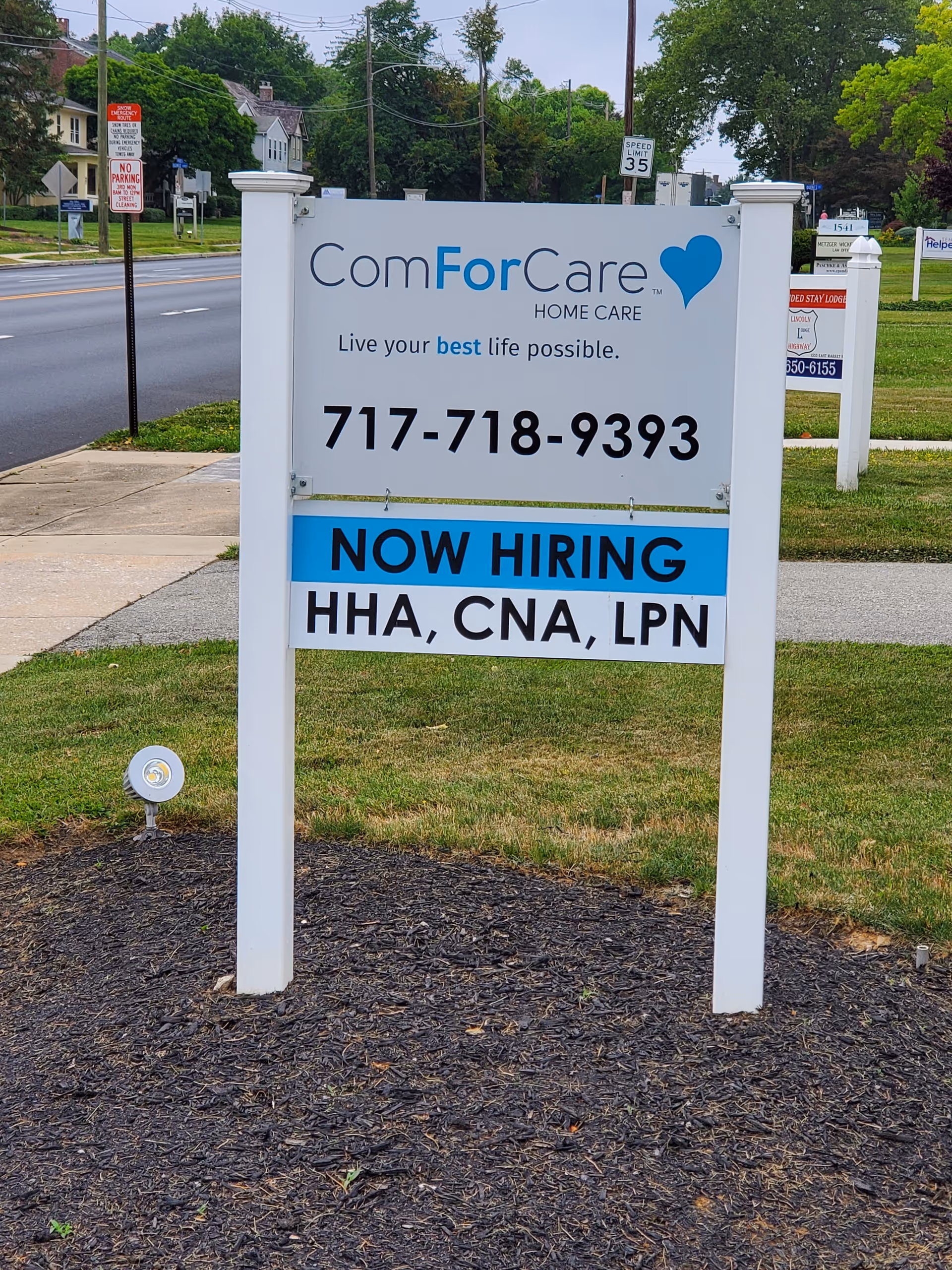 A large white ComForCare sign on posts beside the road with Now Hiring message.