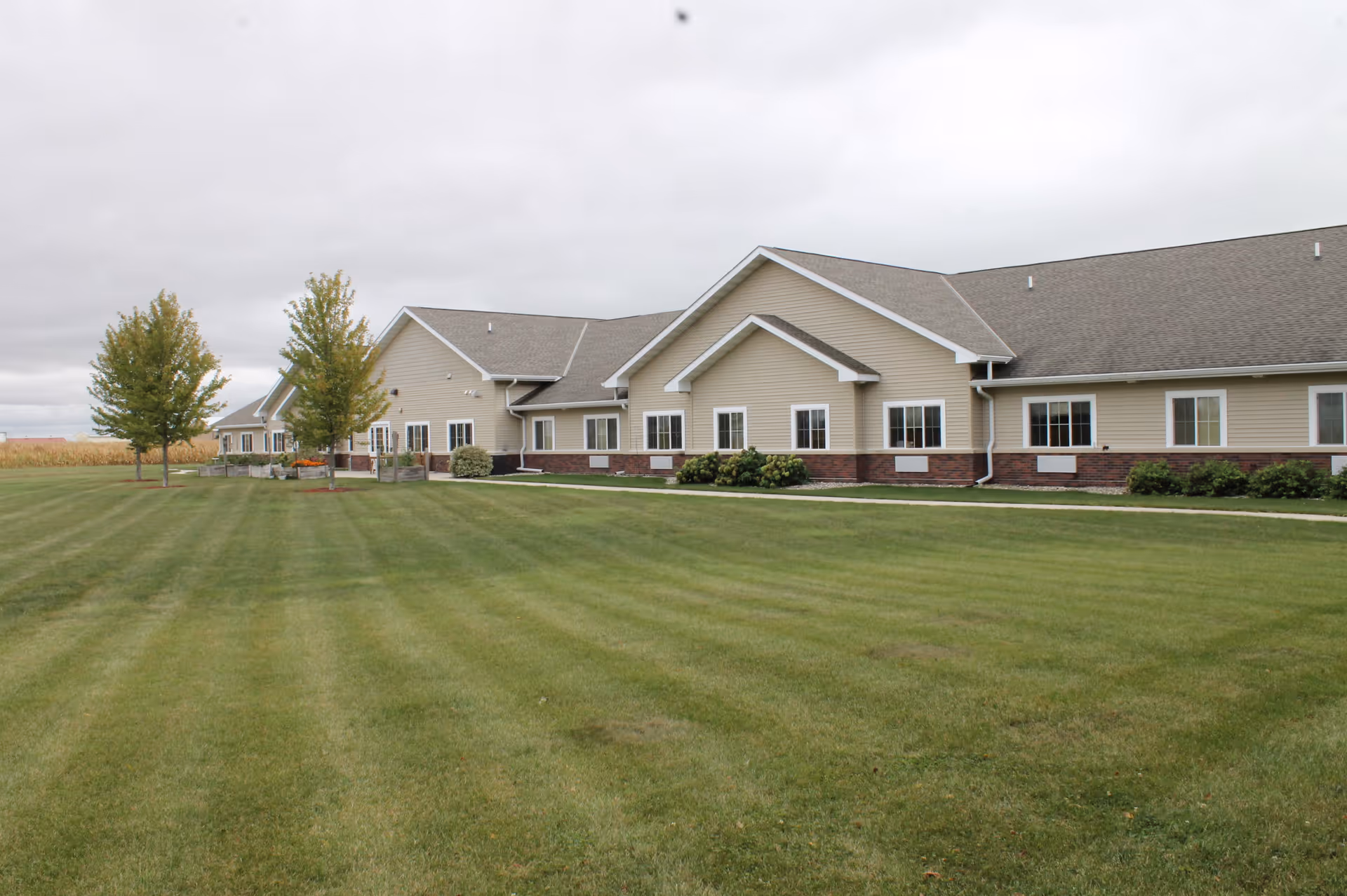 Single-story beige care facility building with a large mowed lawn and a few trees under an overcast sky.
